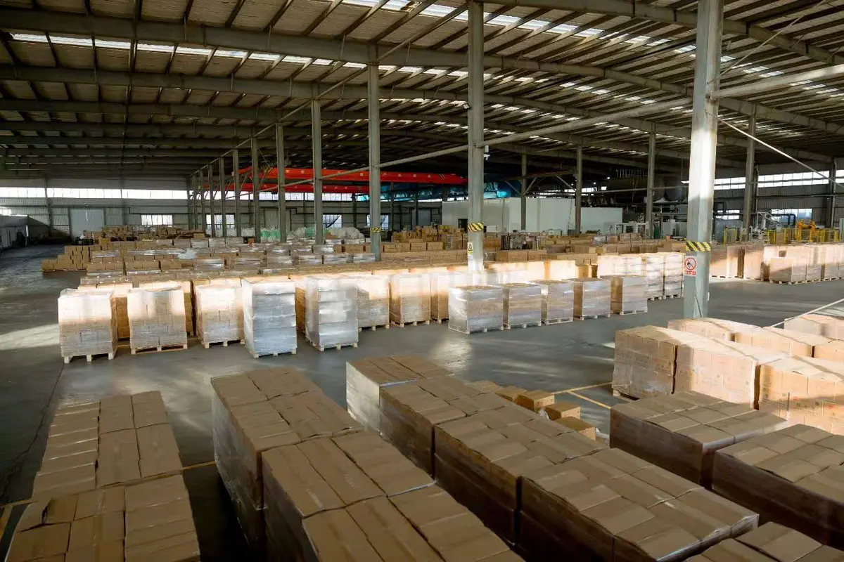 Kho hàng mái cao, các kiện hàng bọc màng xếp thành dãy dài trên sàn, cột chắn sơn vạch vàng đen ở lối đi